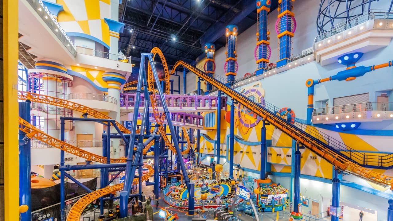 An indoor amusement park features a bright orange roller coaster with blue supports winding through a large, colorful, multi-level building with smaller amusement rides.