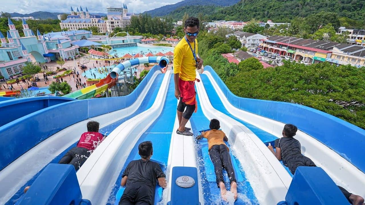 A lifeguard stands between blue-and-white water slides as people slide down towards a water park with castle-like structures and background hills.