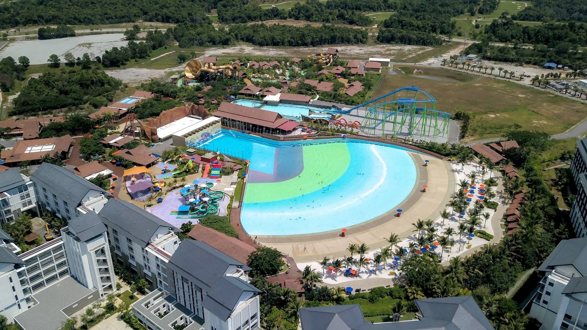An aerial view capturing a water park featuring a large wave pool, various water slides, and resort buildings surrounded by trees and green landscape at Desaru Coast