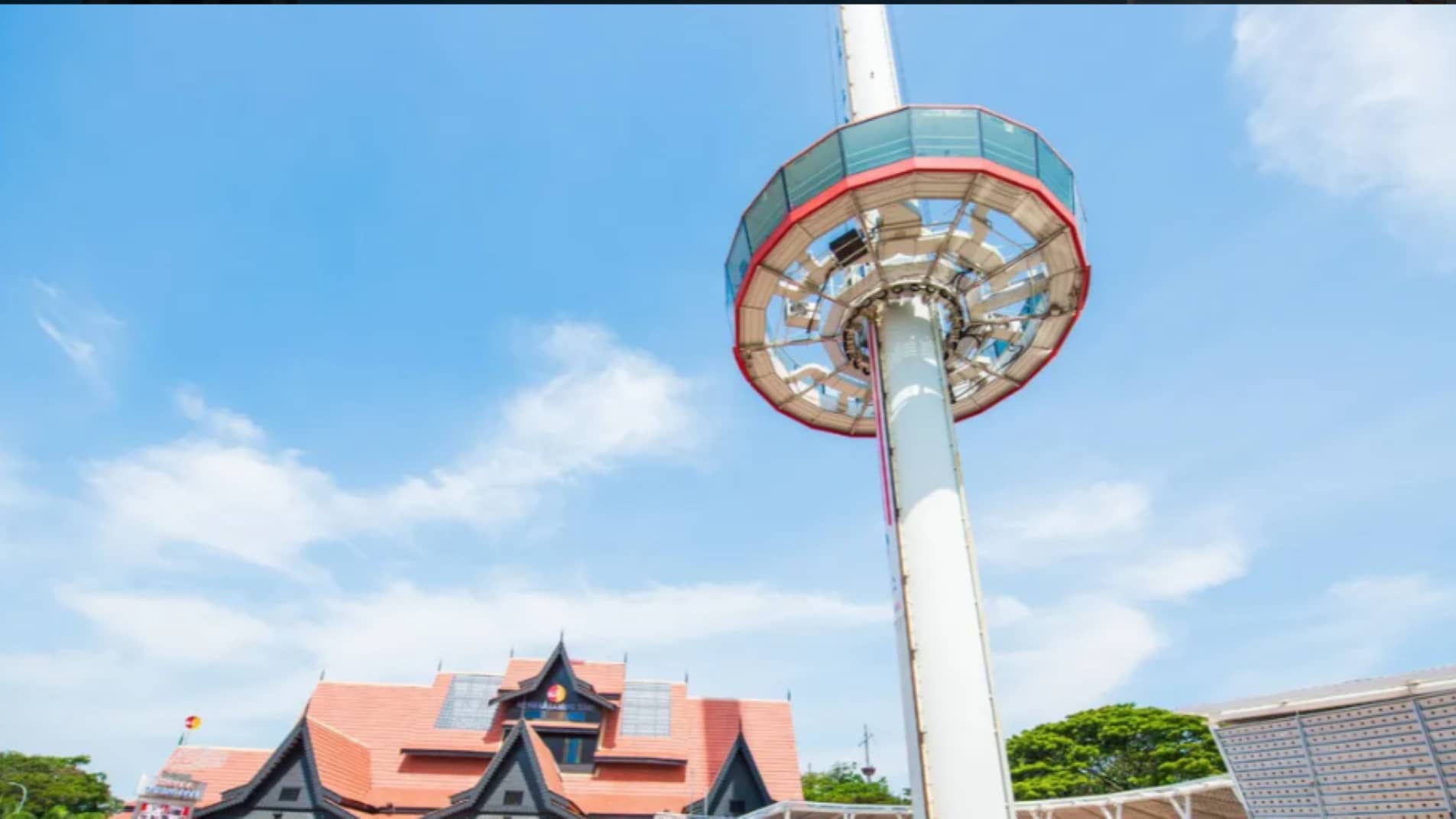A tall observation tower with a circular cabin stands beside a building featuring a red tiled, gabled roof under a blue sky.