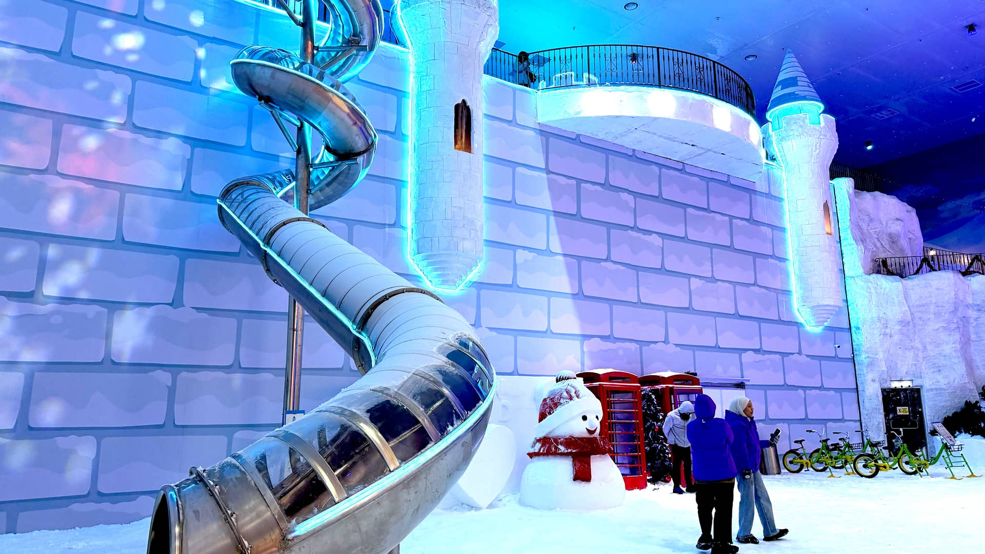 A spiral snow slide, snowman, red telephone booths and people on snow in front of an illuminated castle-themed brick wall with towers.