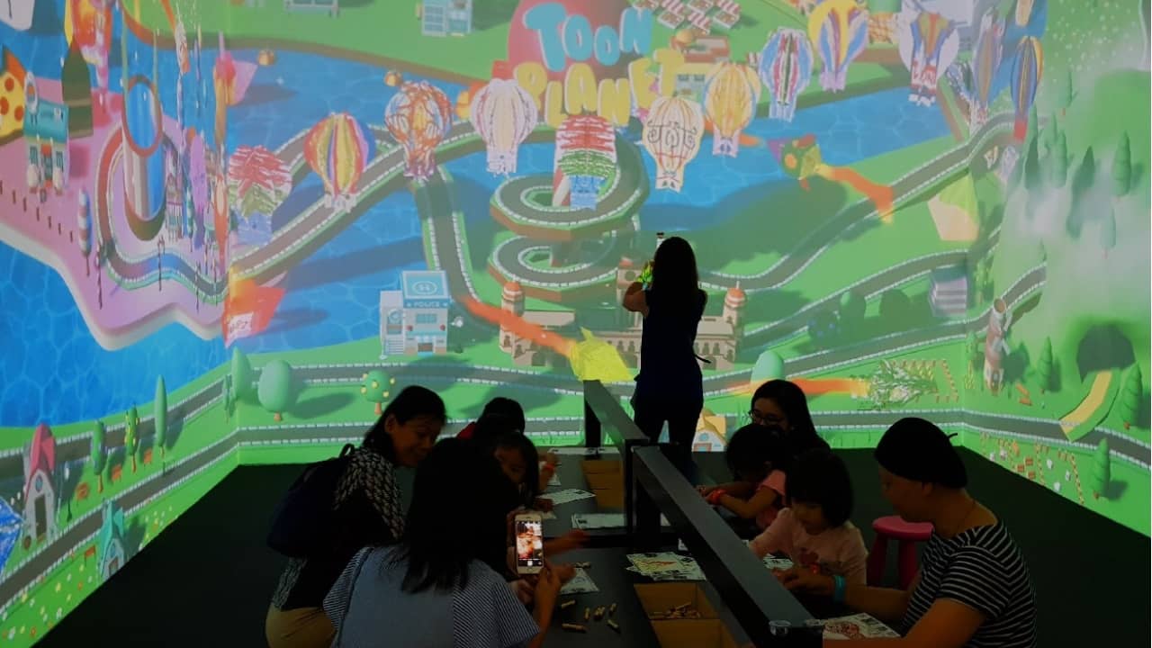 People sit before a large, colorful digital projection of a cartoon landscape featuring buildings, roads, hot air balloons, and the text "Toon Planet."