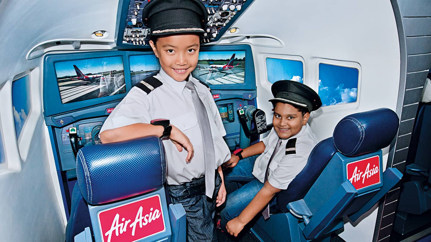 Two children in pilot uniforms sit inside an AirAsia-branded cockpit simulator with screens displaying airplane graphics at KidZania Kuala Lumpur.