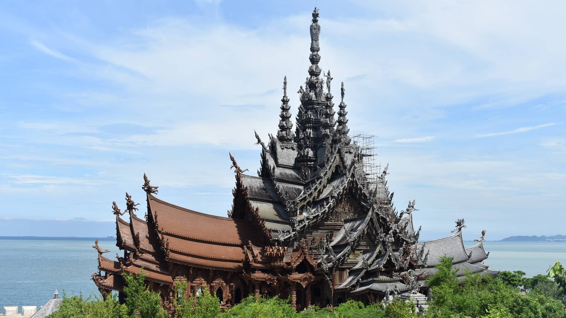 Sanctuary of Truth wooden temple by the water An elaborate, multi-tiered wooden temple stands among green trees beside a vast body of water under a blue sky in Pattaya, Thailand